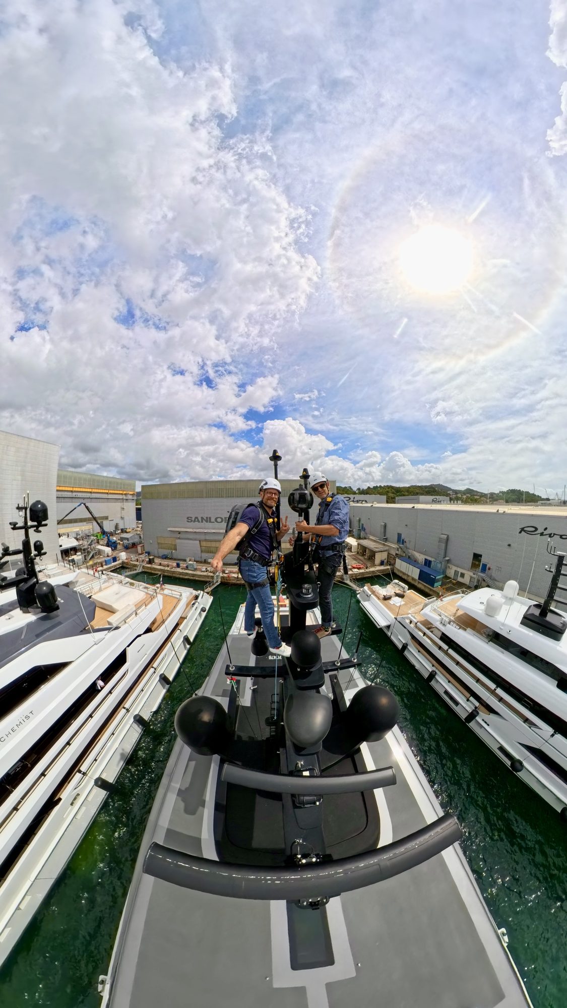 Two Panoblu team members in helmets and harnesses at the mast head of a superyacht, the 360° camera on its pole between them, with the yachts and sheds of the shipyard below.
