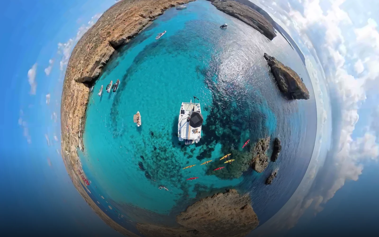 The Blue Lagoon, Comino, seen as a complete 360° little-planet view: the anchored catamaran at the centre, turquoise water, kayakers and limestone cliffs curved around it.